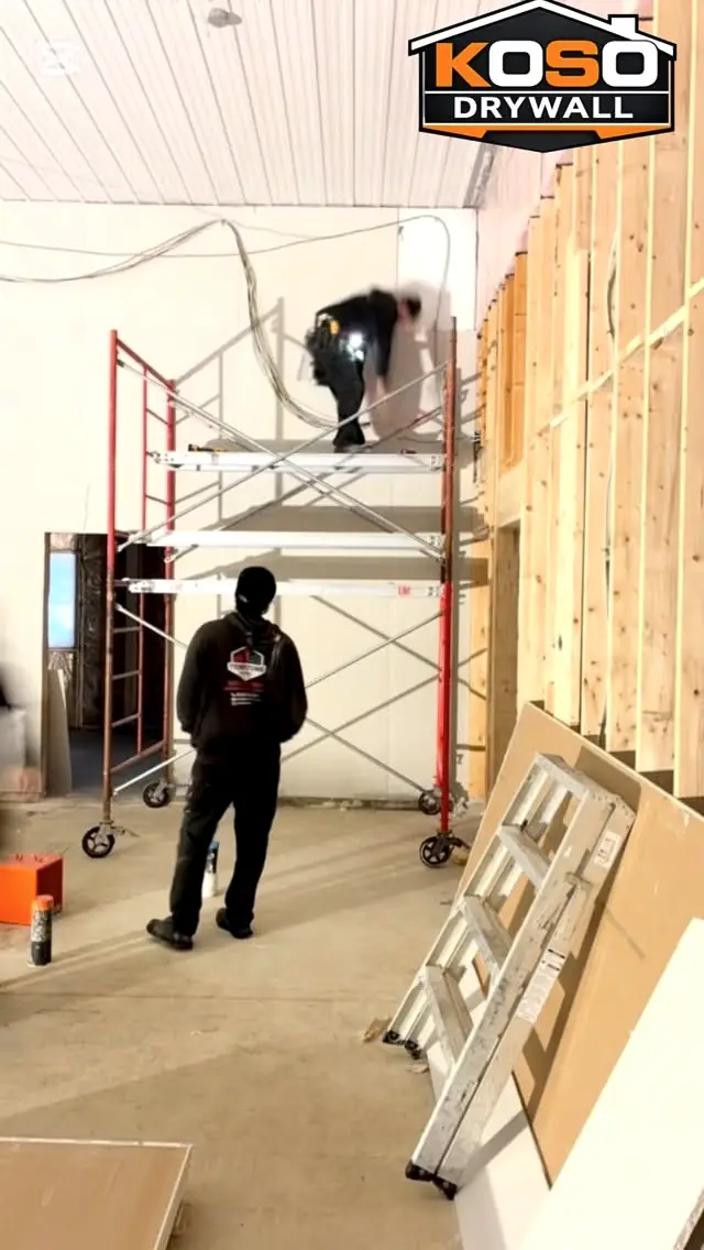 A KOSO crew member on scaffolding hanging drywall in a warehouse, a second crew member watching from the floor
